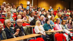 Photo taken of an audience of writers, smiling and laughing, seated in a lecture hall at the University of Lincoln, as part of the Writers' Conference 2025, organised by writing charity and literary development agency Writing East Midlands.